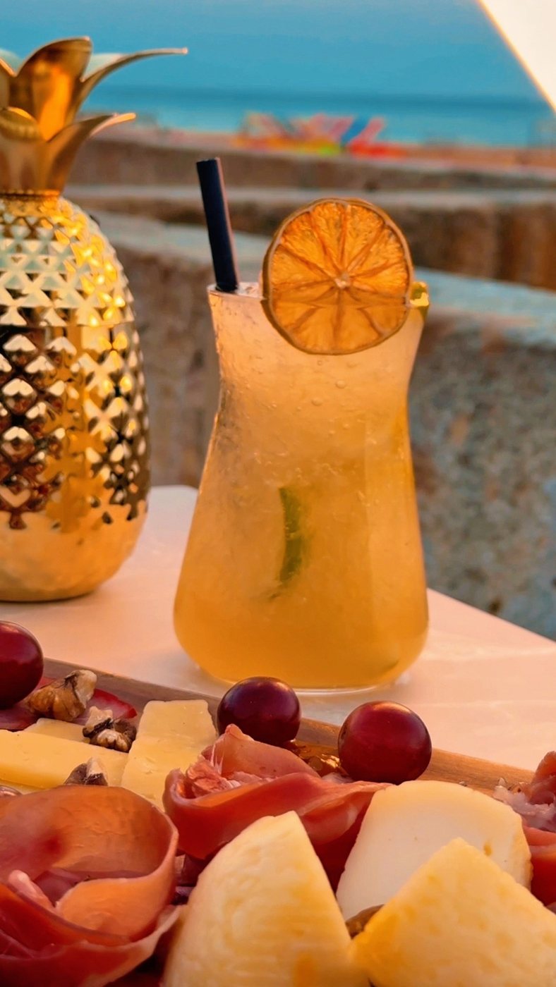 Cocktail e tábua de charcutaria ao pôr do sol, com a praia ao fundo